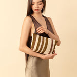 Artisan hand woven Cristina Cosmetic Pouch in Forest. A model holds the pouch in one arm and holds the leather zipper pull with her hand. The forest pattern has vertical olive green and beige stripes.
