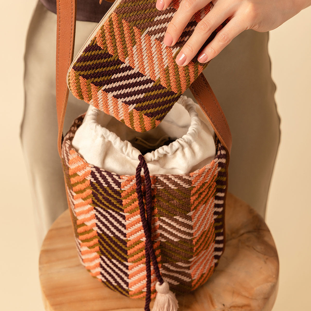 Hand woven artisan Betty Cylinder Crossbody and Mini Lily Wristlet in Moss Weave. A model places the Mini Lily Wristlet inside the Betty Cylinder. The Betty Cylinder has a drawstring bag with peach tassels. The Moss Weave is an olive green and orange plaid.