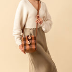 Hand woven artisan Betty Cylinder Crossbody in Moss Weave. The Moss Weave is an olive green and orange plaid. It has an adjustable leather strap. A model wears the Betty Cylinder crossed over her torso.