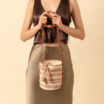 Hand woven artisan Betty Cylinder Crossbody in Snow. A model holds the leather straps wraped around her hands. The Crossbody has horizontal pale pink and beige stripes and pale pink tassels.