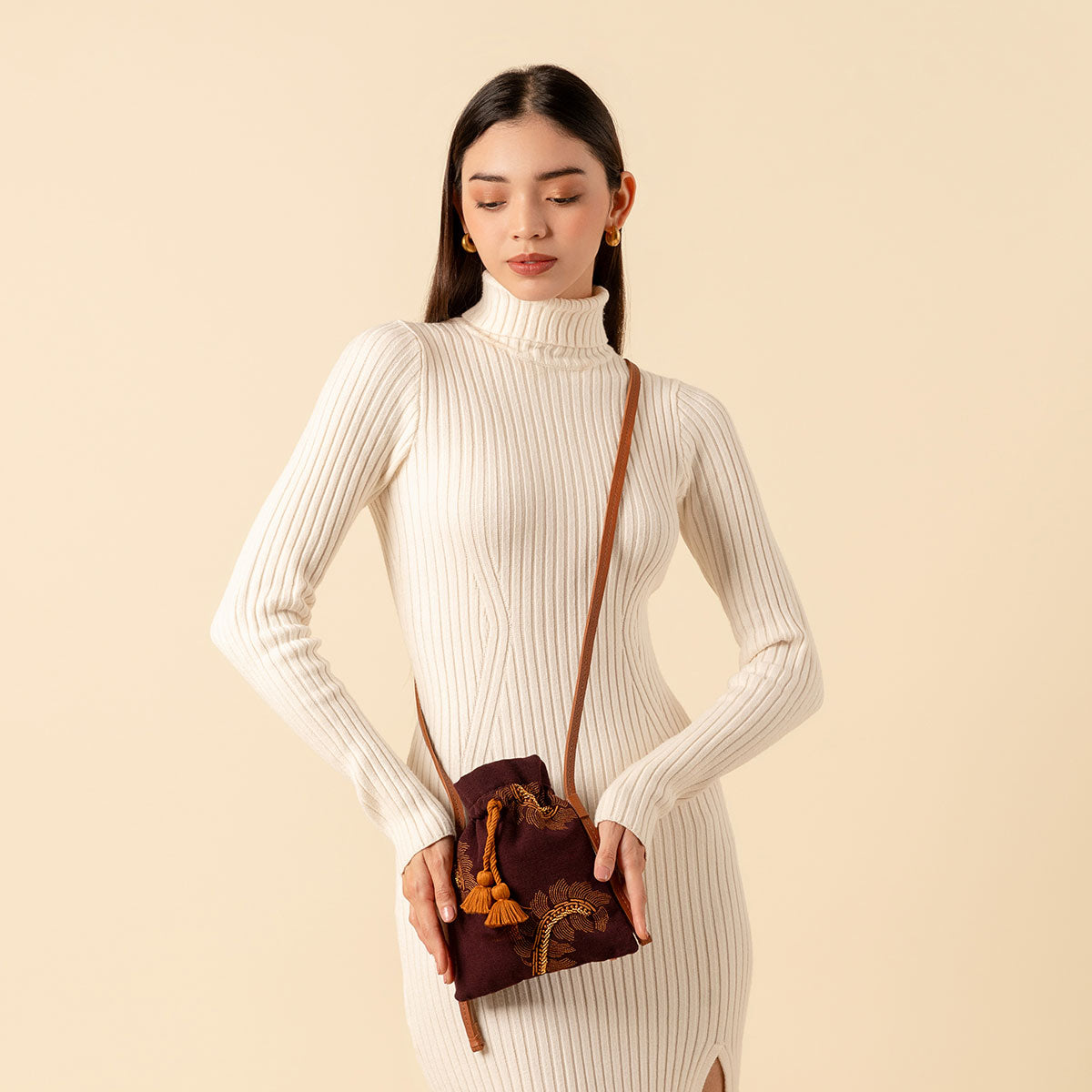 Hand woven artisan Eloisa Small Crossbody in Season Sparkle. A model wears the crossbody around her torso. The Eloisa Crossbody has embroidered detailing, a drawstring shape with orange tassels, and a leather strap.