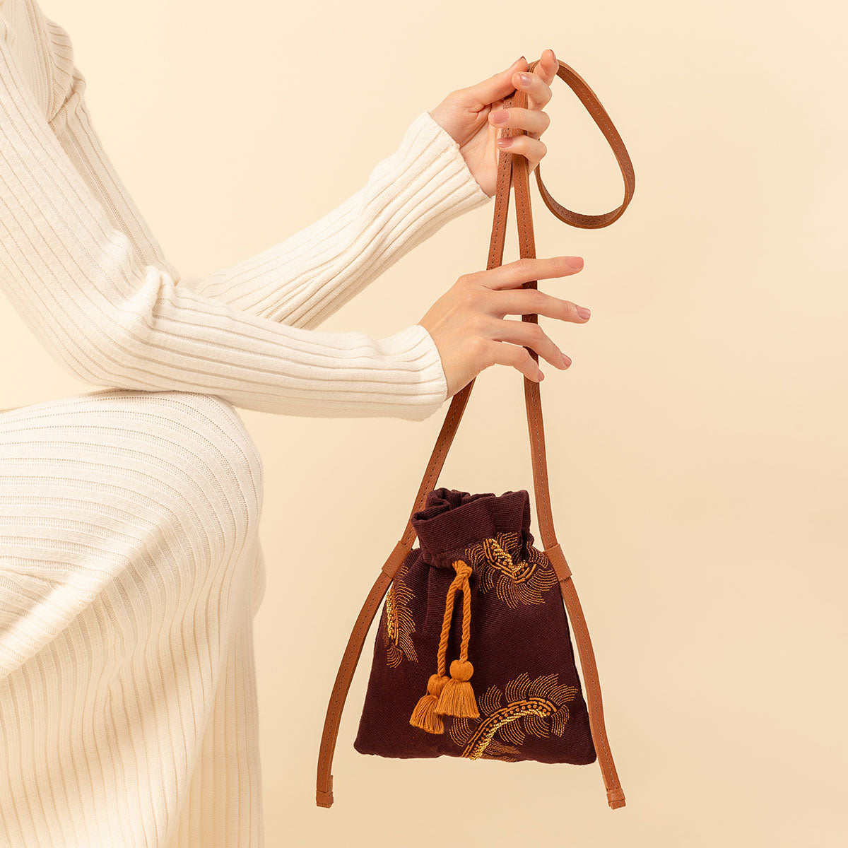 Hand woven artisan Eloisa Small Crossbody in Season Sparkle. A model holds the small crossbody. It has a leather handle, a drawstring shape with orange tassels, and embroidered detailing.