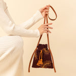 Hand woven artisan Eloisa Small Crossbody in Season Sparkle. A model holds the small crossbody. It has a leather handle, a drawstring shape with orange tassels, and embroidered detailing.