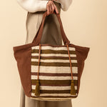 Hand woven artisan Rosa Tote in Forest pattern. The forest pattern has olive green and beige horizontal stripes with triangular brown finishes and handles. It has two tassels attached to the handles. A model holds the tote in one hand.
