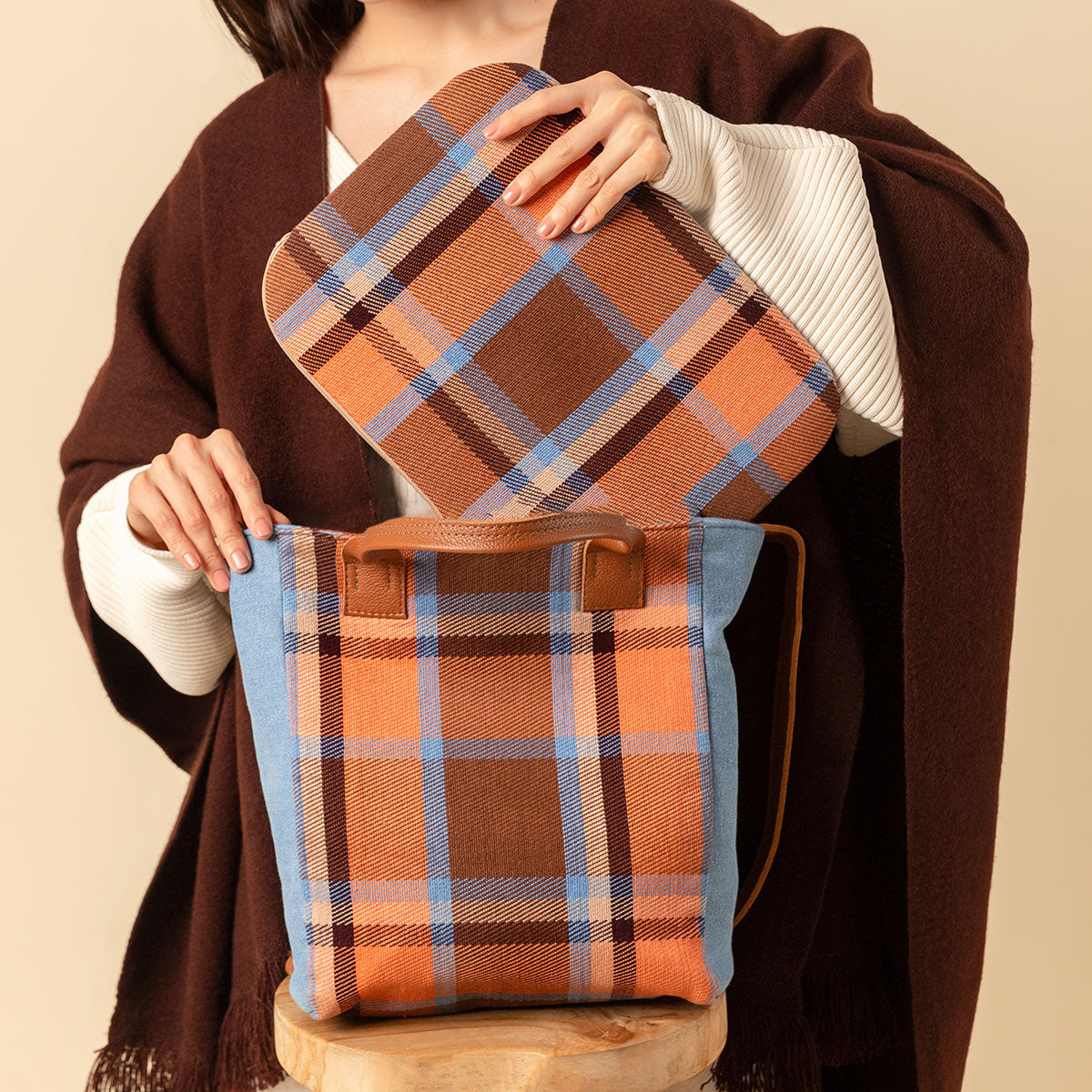 Featured is the hand woven artisan Ana Tote and Elenita Laptop Case both in the Brickstone pattern. A model puts the laptop case into the Ana tote. She is wearing a brown coat over a white sweater. The Ana tote sits on a wooden stool. The background color is light tan.