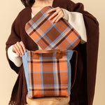 Featured is the hand woven artisan Ana Tote and Elenita Laptop Case both in the Brickstone pattern. A model puts the laptop case into the Ana tote. She is wearing a brown coat over a white sweater. The Ana tote sits on a wooden stool. The background color is light tan.