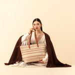 Hand woven artisan Estella Tote in Snow pattern. A model holds the Estella tote and sits on the ground, looking at the camera. A brown coat is draped over her. The tote has horizontal beige and pale pink stripes.