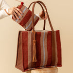 Hand woven artisan Irma Tote and Mini Lily Wrislet in Chestnut color. A model is shown putting the wristlet into a tote. The Chestnut pattern has vertical burgundy, tan, and beige stripes. It has brown solid color sides and handles.