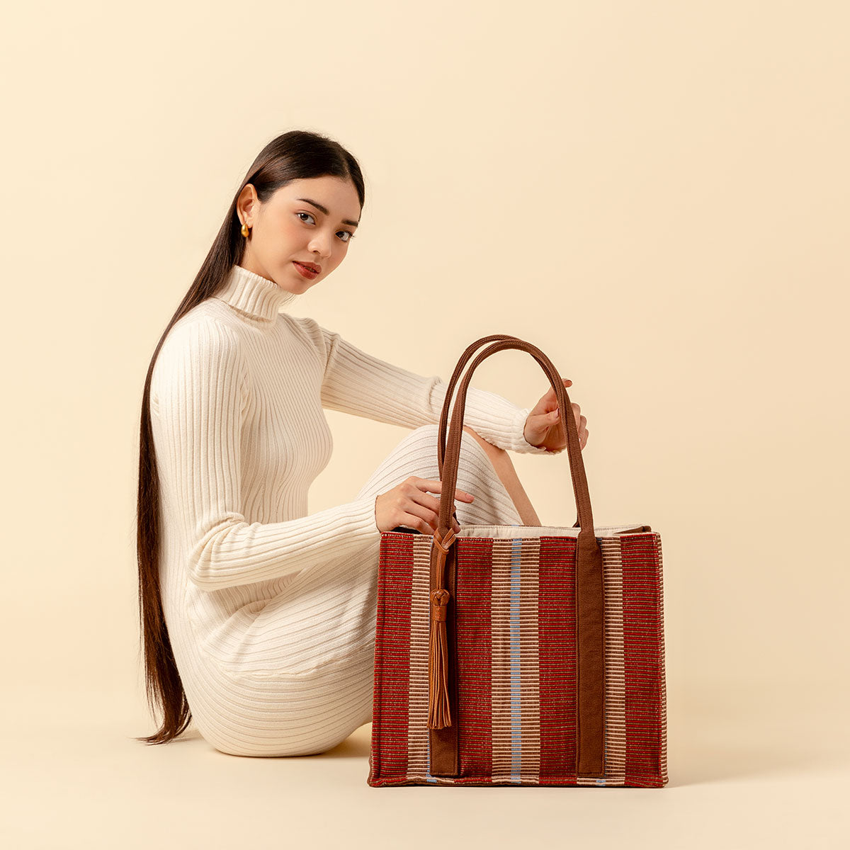 Hand woven artisan Irma Tote in Chestnut pattern. A model sits and displays the Irma Tote. The chesnut pattern has burgundy and tan stripes. The tote has brown handles with a leather tassel attached. The model looks at the camera over a cream yellow colored background.