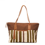 Hand woven artisan Soledad Weekender in Forest pattern. The front of the bag is shown with a horizontal brown fabric, vertical beige and green stripes, leather handles, a tassel, and a leather zipper pull to the left.
