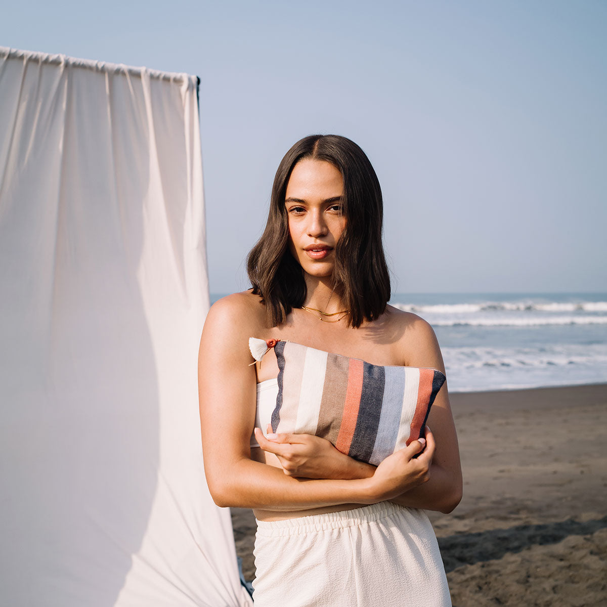 A model holds the Cristina Pouch in Blue Canyon. She stands in front of a white tent on the beach.