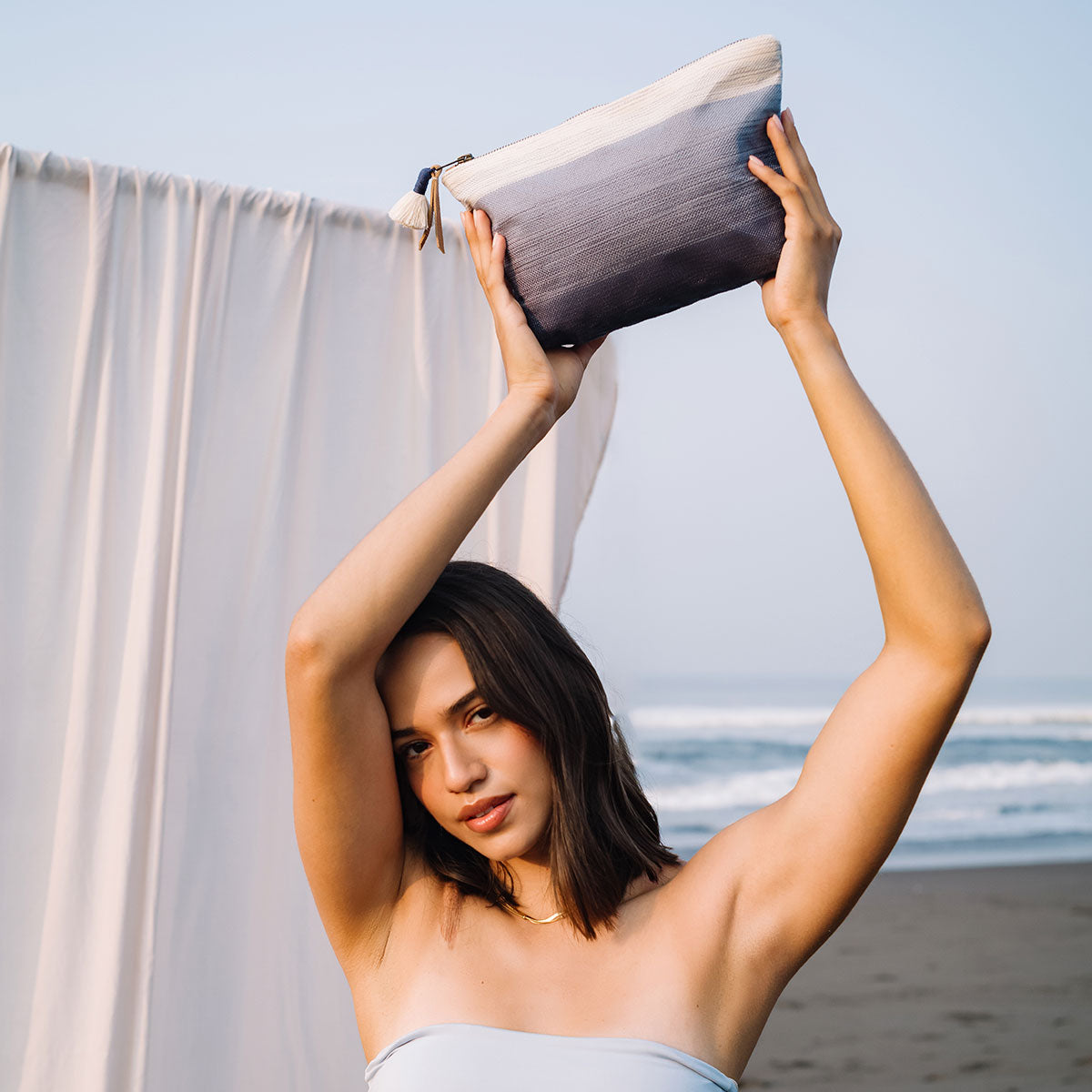A model holds the Cristina Pouch in Ocean Breeze over her head. She stands in front of a white tent on the beach.
