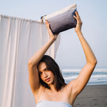 A model holds the Cristina Pouch in Ocean Breeze over her head. She stands in front of a white tent on the beach.
