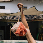 A model holds the Dulce Wristlet in Desert Sands. The background set is a courtyard with a pool, furniture, and patio covering.