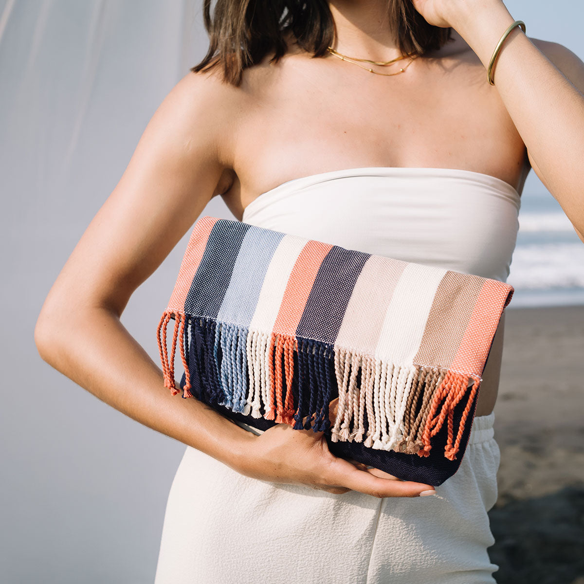 A model holds the Margarita Clutch in Blue Canyon. The background is a white tent on the beach.