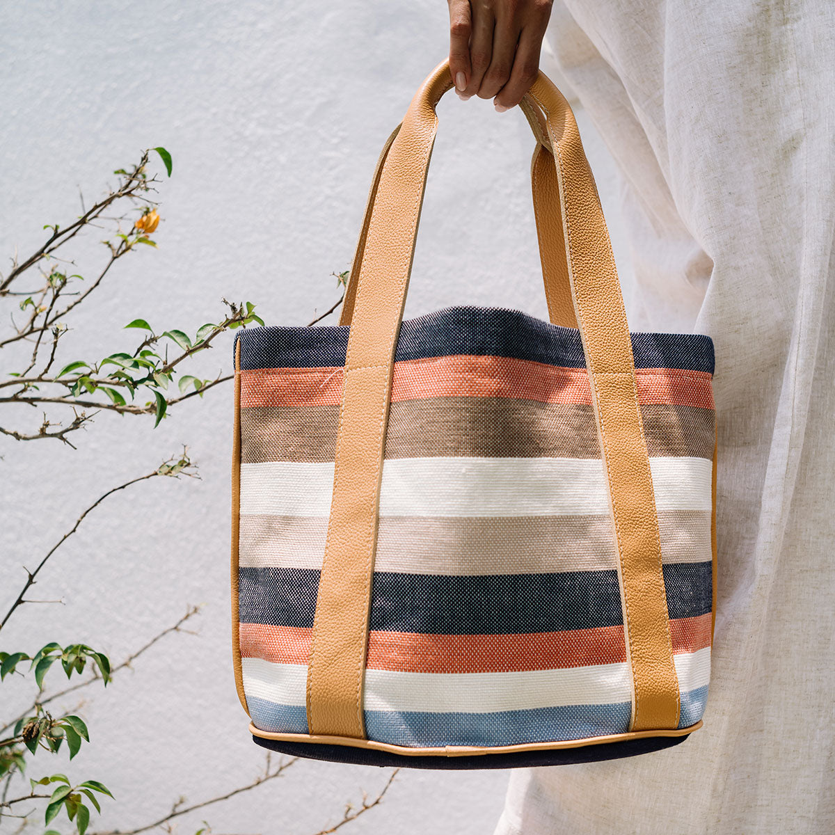 A model holds the Magali Tote in Blue Canyon. The background is a white wall and plants.