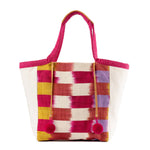The front of the artisan hand woven Rosa Tote in Raspberry Paleta. It has a flame stitch red, orange, yellow, purple, and white stripes. It has two pink pompoms attached to leather cords.