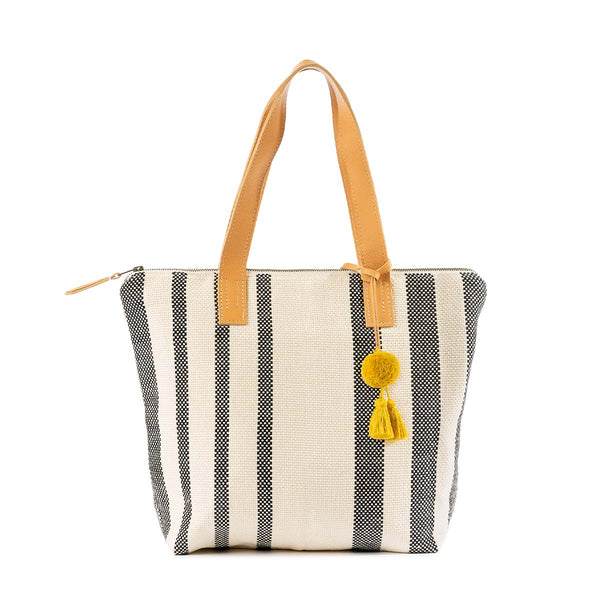 The front of the hand woven artisan Angela Tote in Tourmaline pattern. It has grey and white vertical stripes, leather handles, and a yellow pompom-tassel combination.