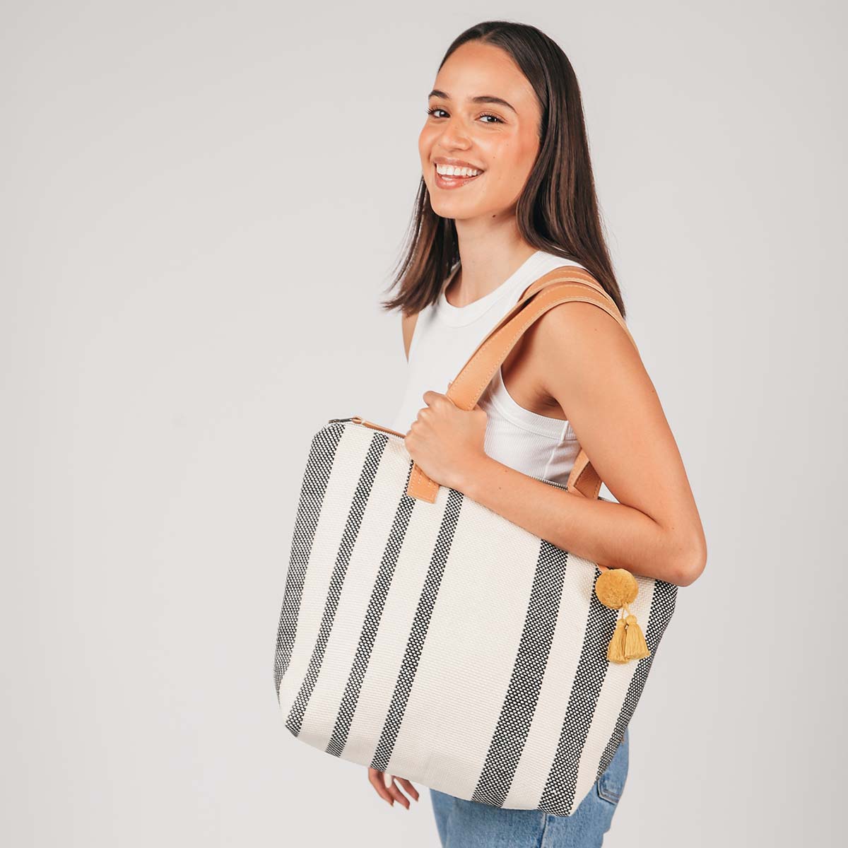 A model holds the Angela Tote in Tourmaline pattern over her shoulder. It has grey and white vertical stripes, leather handles, and a yellow pompom-tassel combination.