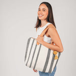 A model holds the Angela Tote in Tourmaline pattern over her shoulder. It has grey and white vertical stripes, leather handles, and a yellow pompom-tassel combination.