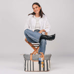 A model sits and poses near the Soledad Weekender in Tourmaline pattern. It has grey and white vertical stripes, leather handles, and a yellow pompom-tassel combination. The Soledad Weekender has an Ines Luggage Tag in 90's Denim.