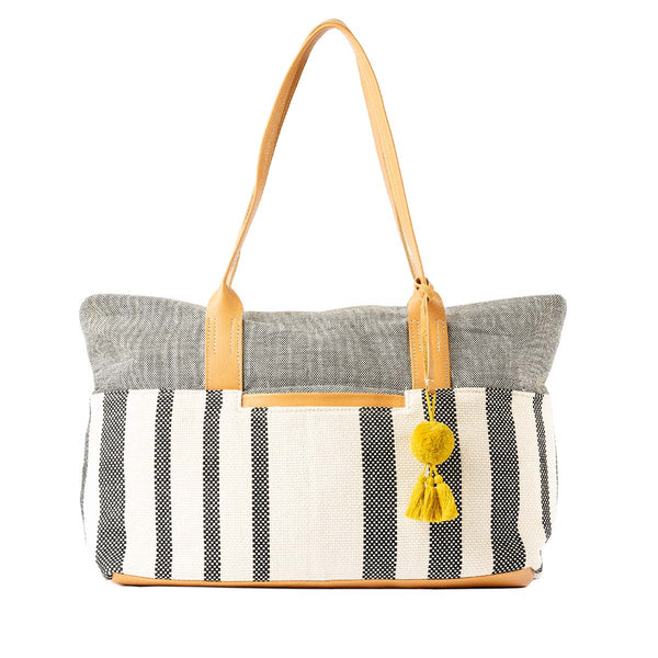 The front of the Soledad Weekender in Tourmaline pattern. It has grey and white vertical stripes, leather handles, and a yellow pompom-tassel combination.