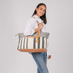A model holds the Soledad Weekender in Tourmaline pattern over her shoulder. It has grey and white vertical stripes, leather handles, and a yellow pompom-tassel combination.