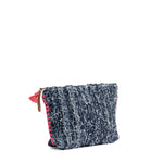 A side angle of the artisan hand woven Cristina Cosmetic Pouch in Denimology. The Cristina Pouch has strips of denim woven together with thick coral pink visible side stitches. It has a coral pink mini tassel and a leather zipper pull.