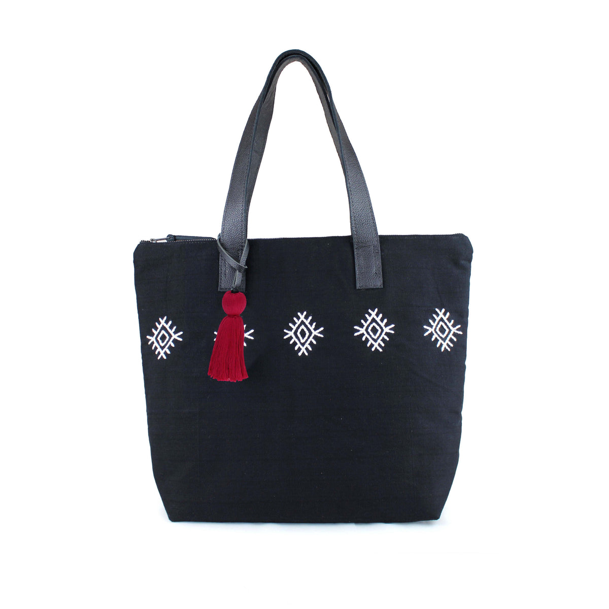 Front view of the Angela Tote with black and white diamond embroidery. It has a red tassel and dark leather handles.