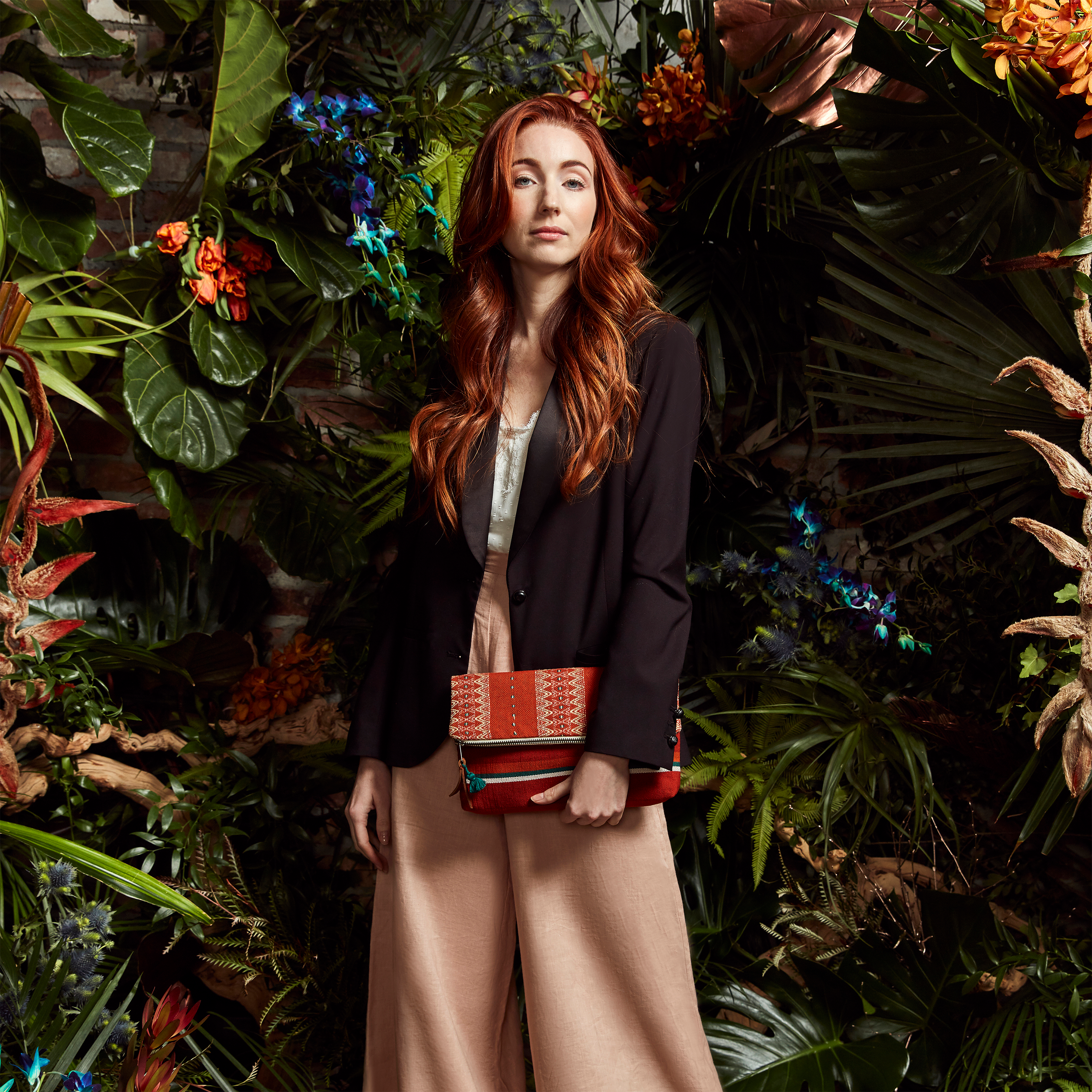 A model stands in front of tropical plants and looks at the camera. She holds the hand woven artisan Celia Clutch in Ginger pattern in one hand.