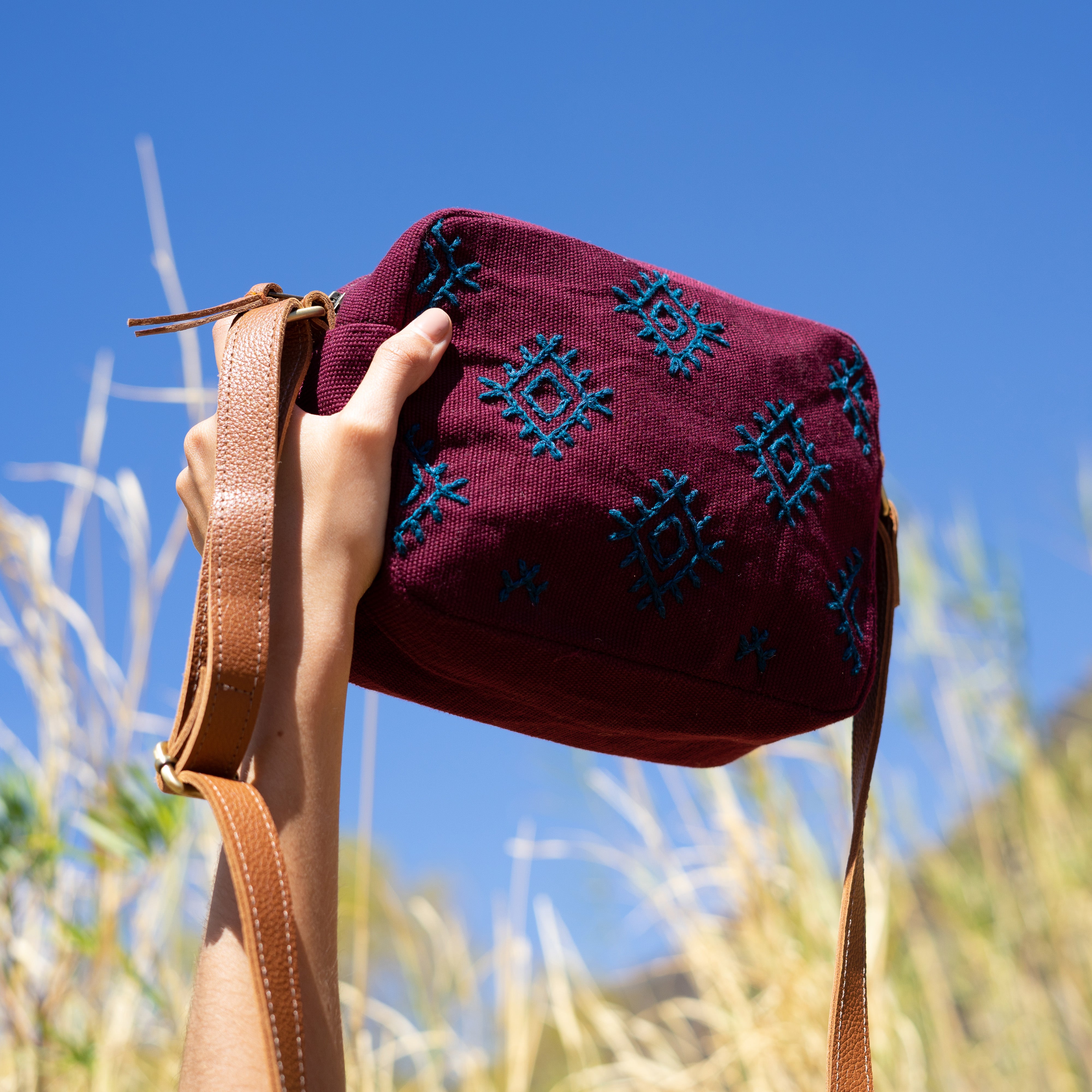 A model holds up the hand woven artisan Juana Crossbody bag in Mountain Dusk. The photo is taken outside in a grassy field.