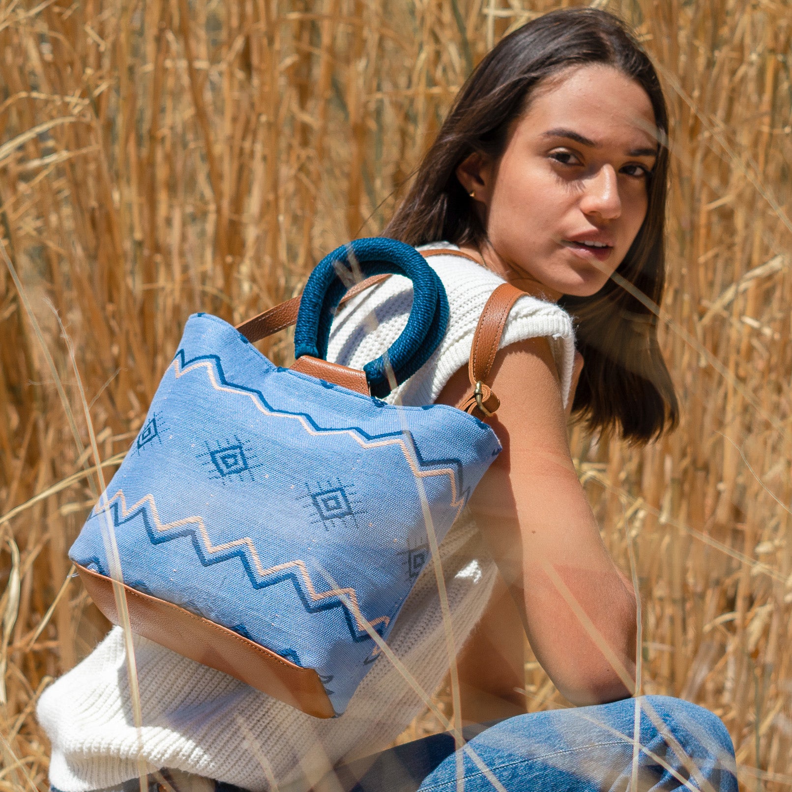 A model poses outside in a wheat field and wears the hand woven artisan Dalila Midi Tote in Mountain Blue over her shoulder. 