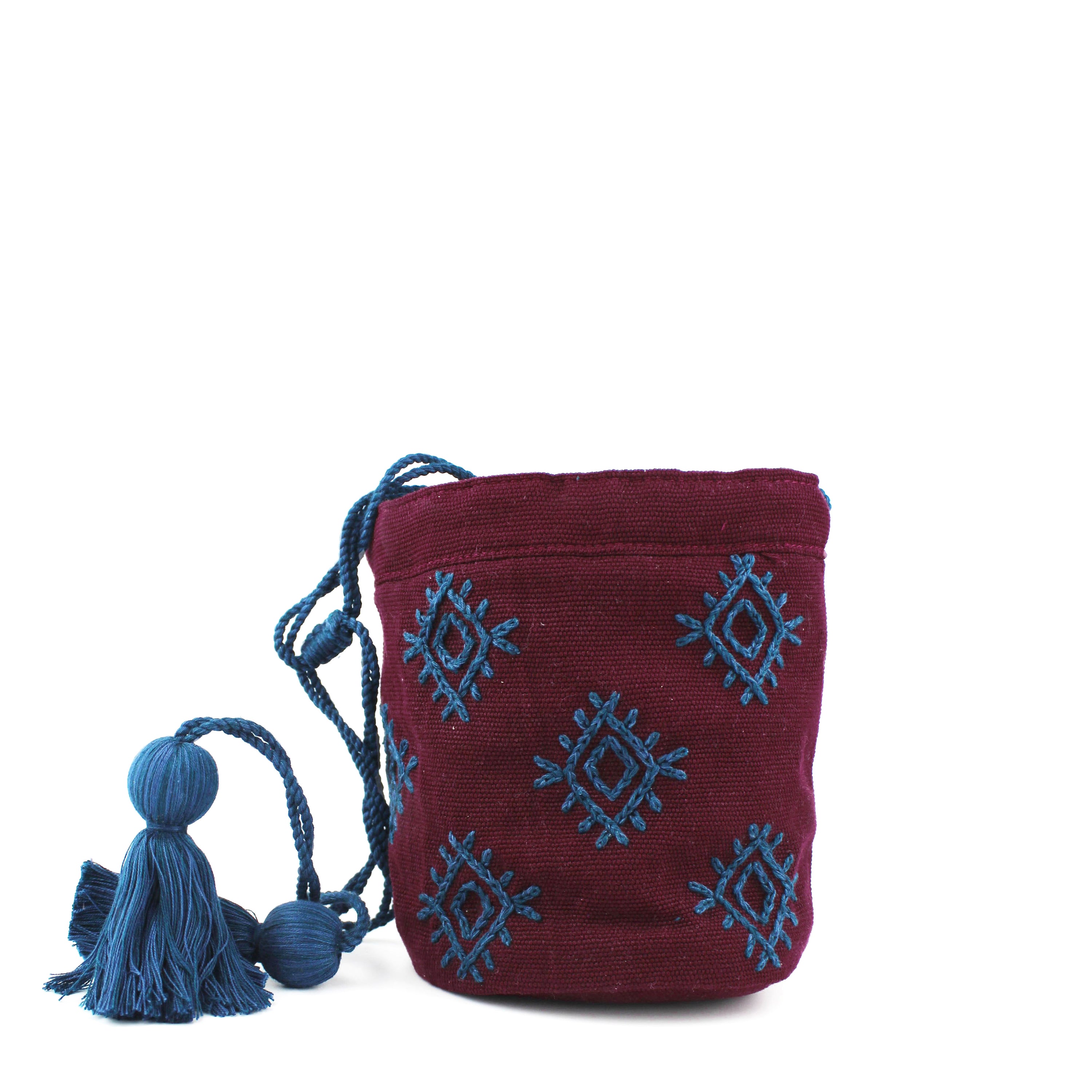 A view of the artisan hand woven Hermelinda Crossbody bag in Mountain Dusk. It has a cylinder shape when opened, with blue drawstrings and tassels threaded inside the hem. The Mountain Dusk pattern has blue embroidered diamond shapes over a maroon background.