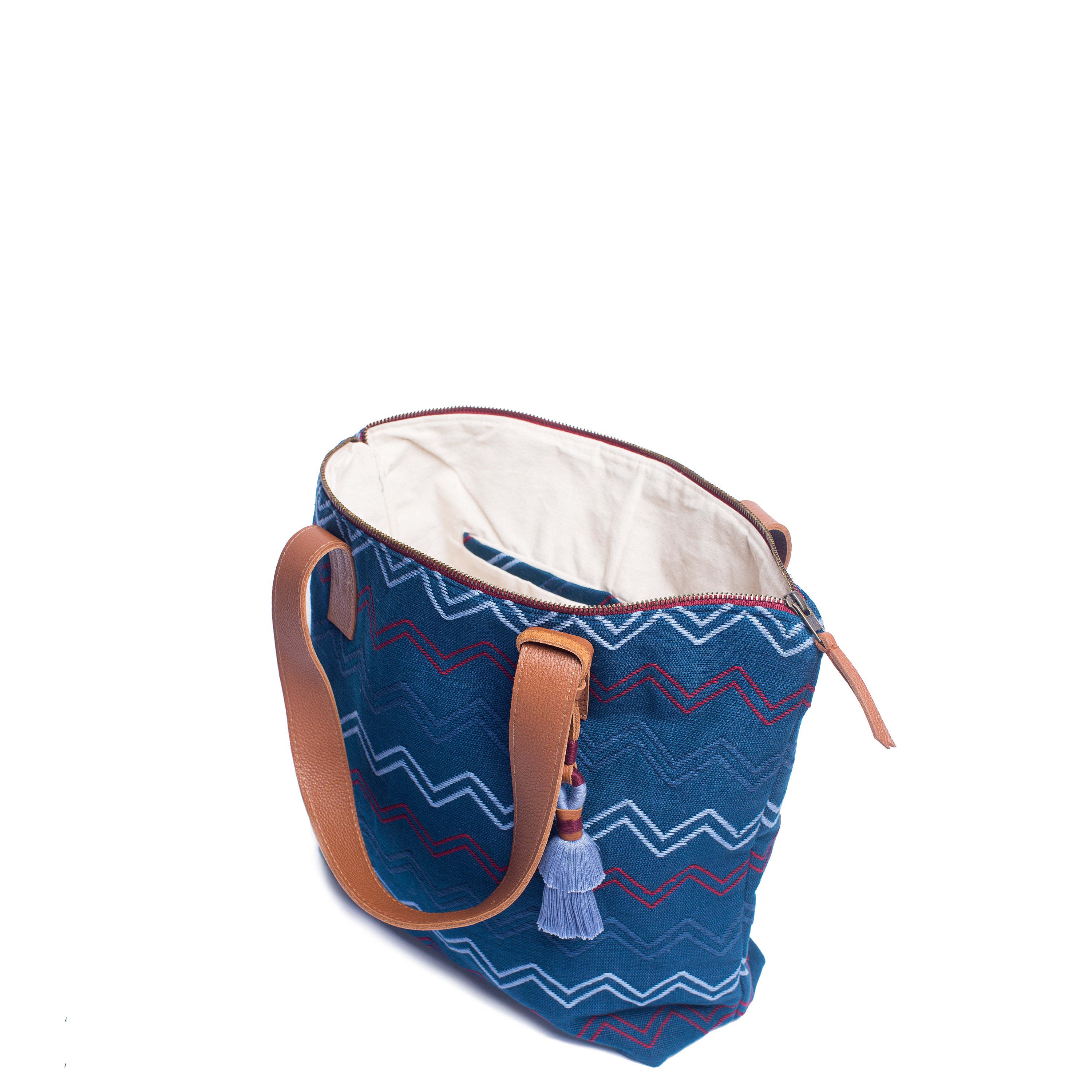 The hand woven artisan Angela Tote is unzipped and open. The interior has a white lining and blue hem. It has horizontal zigzag stripes in red, light blue, and blue over a blue background. It has a blue tassel and leather handles. 