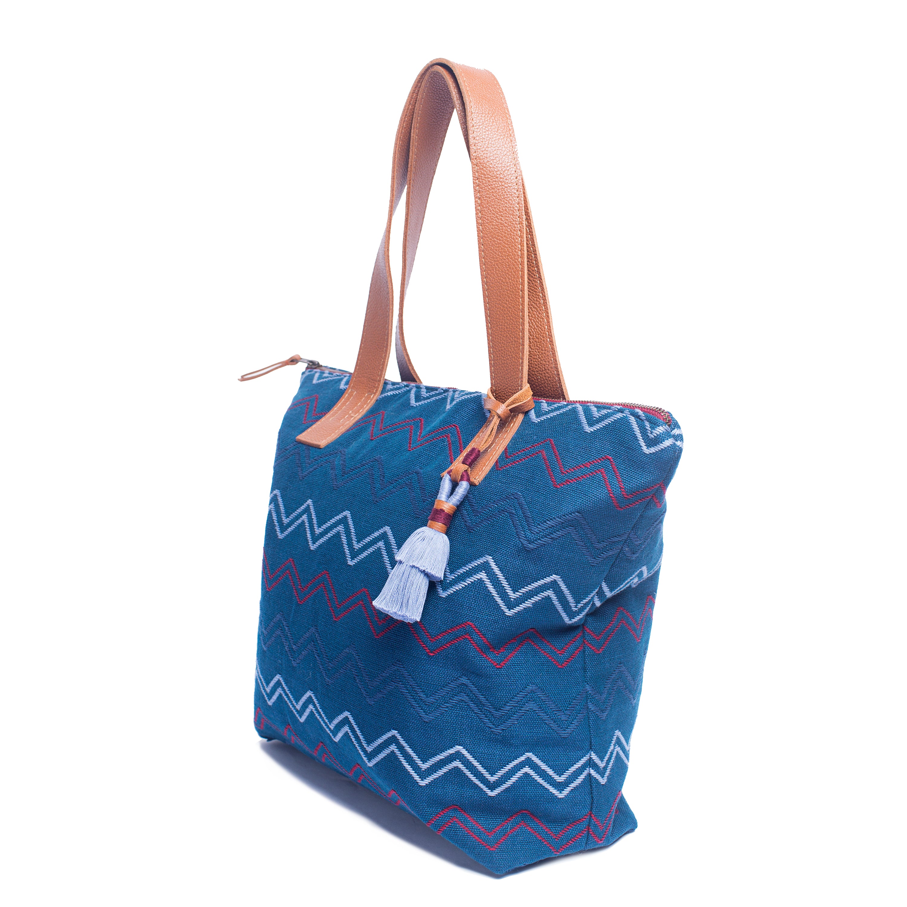 The right-sided angle of the Angela Handwoven Tote in Lake Ripple pattern. It has horizontal zigzag stripes in red, light blue, and blue over a blue background. It has a blue tassel and leather handles.