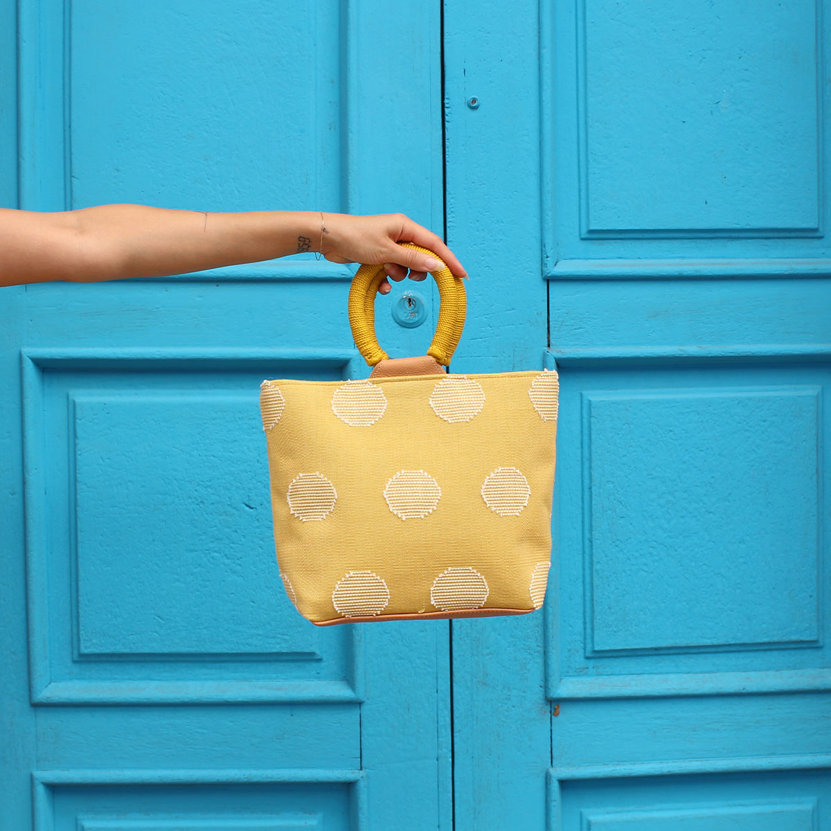 A model holds the hand woven artisan Dalila Midi Tote in Sunset Yellow over a blue painted door.