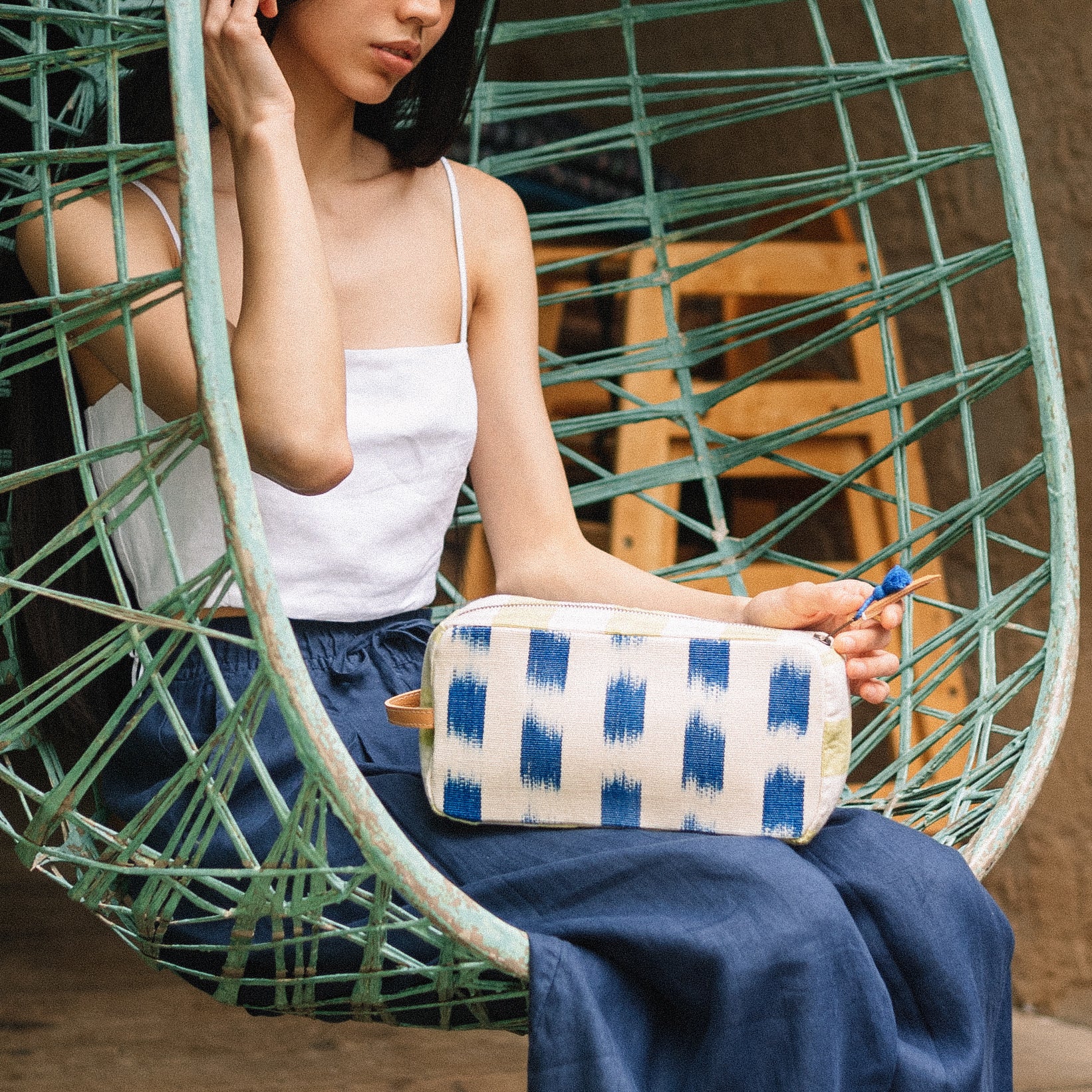 A model sits in a canopy chair and holds the hand woven artisan Edna Dopp Kit on her lap. The Edna has the Brustrokes pattern.