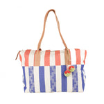 Hand woven artisan Soledad Weekender in Blueberry. The pattern has blue geometric stripes and orange stripes on the top fabric. It has leather handles. Attached are two orange and green pompoms with leather straps.