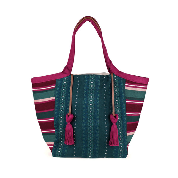 Artisan Rosa Handwoven Tote in Deep Forest Mulberry. The center fabric has a dark green and blue geometric pattern. The sides have magenta, maroon, green, and white horizontal stripes. It has two magenta tassels attached to leather cords. It has leather detailing on the handles.