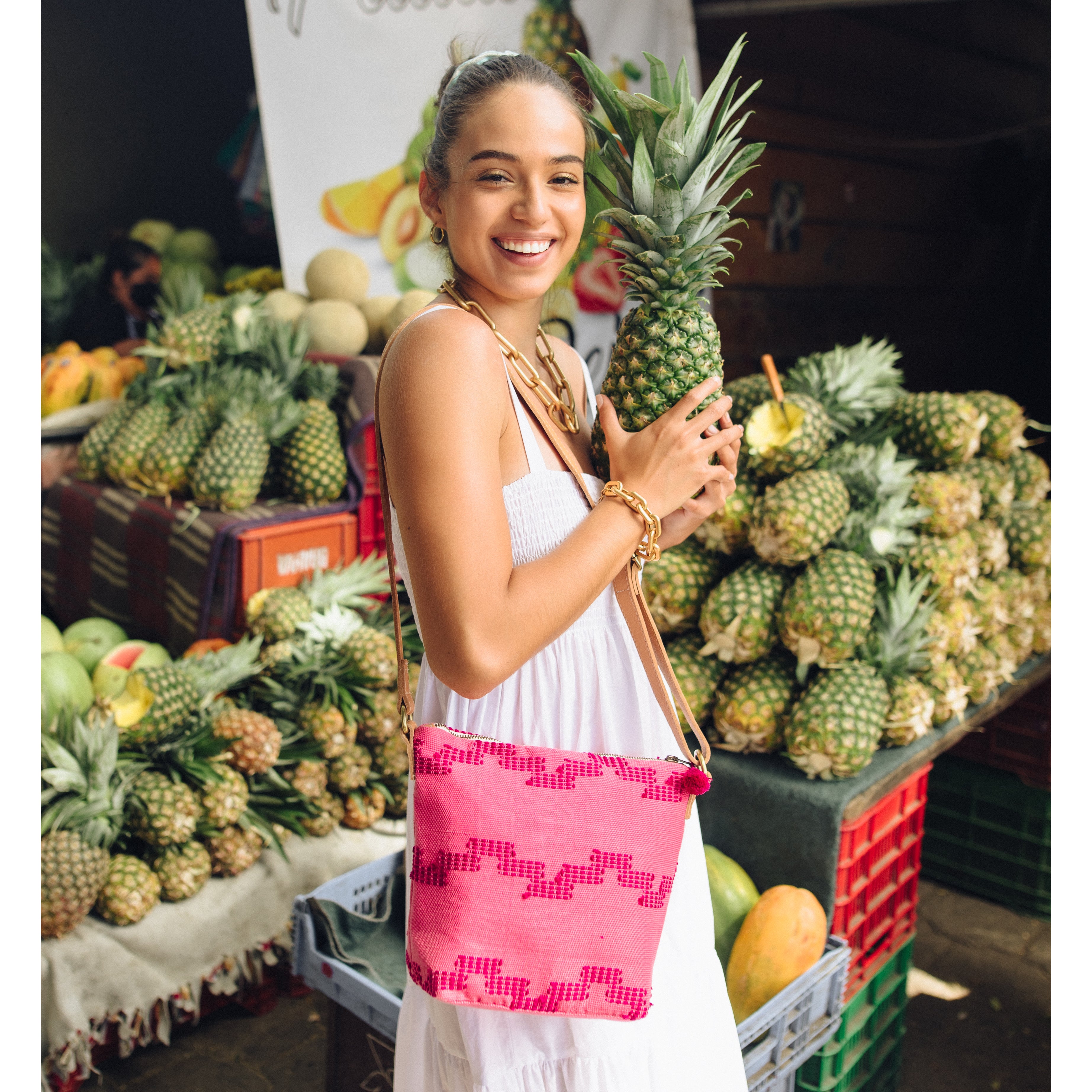 A full photo of a model wearing the hand woven artisan Mini Lidia Crossbody in Pitaya style. The model poses in front of a fruit vendor and holds a pineapple.