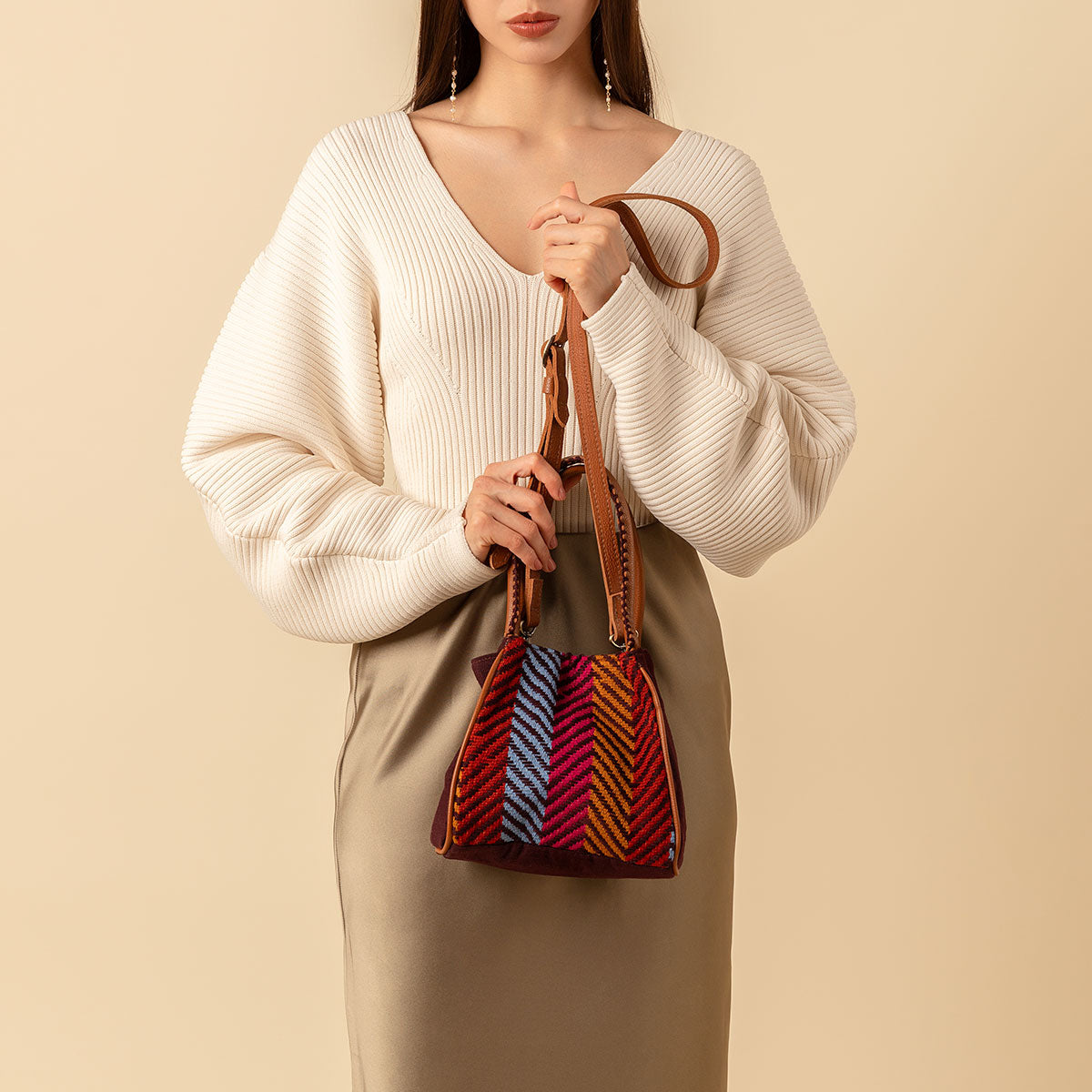 A model holds the leather straps of the Flora Petite Tote in Winter Berry in both hands. The winter berry has vertical stripes in red, blue, and yellow ochre. It has adjustable leather straps.
