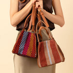 A model holds two Flora Petite Totes in Winter Berry and Chestnut. The Winter Berry has ochre yellow, blue, and red stripes. The Chestnut has vertical brown and tan stripes.