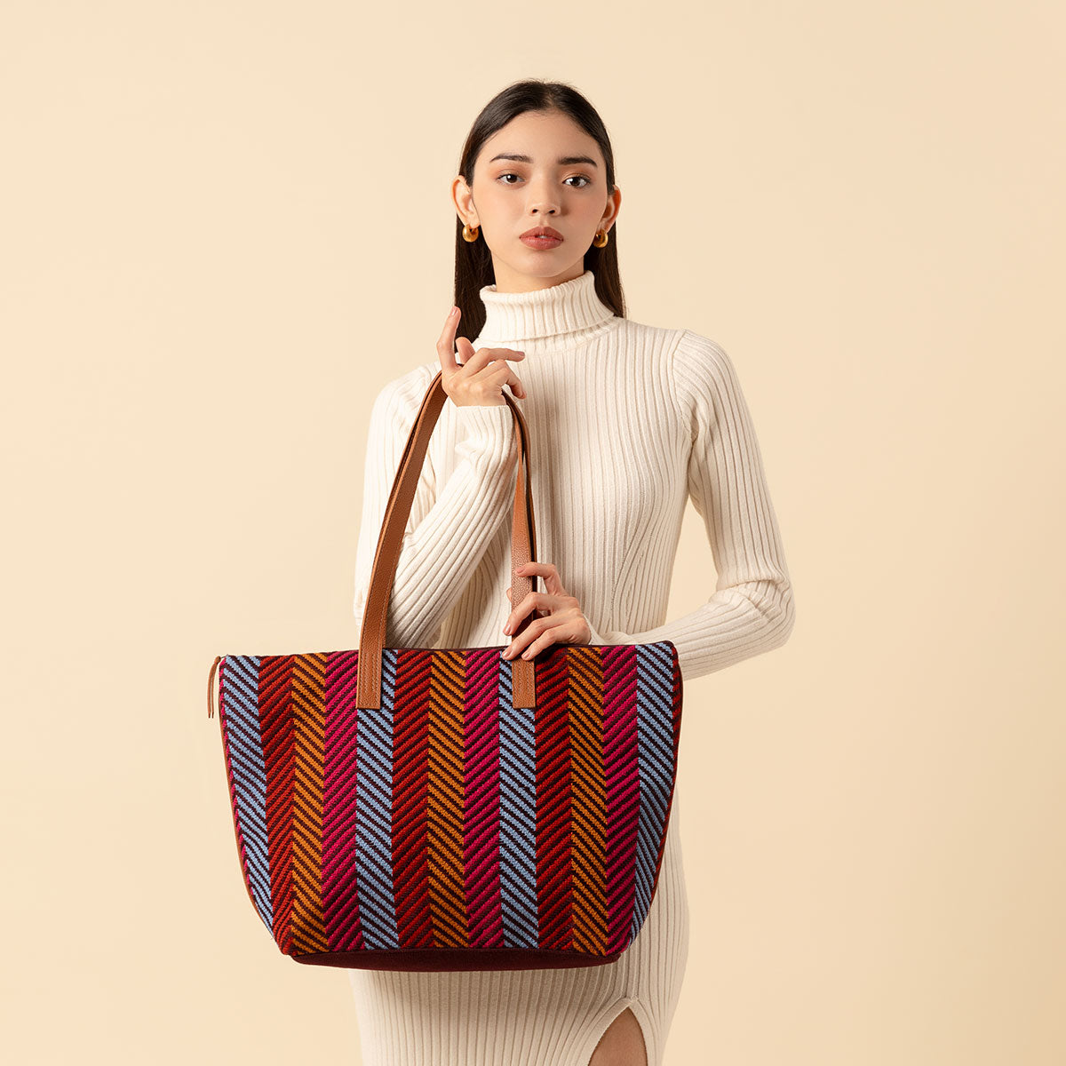 Hand woven artisan Estella Tote in Winter Berry pattern. A model holds the tote and looks at the camera. The winter berry pattern has blue, red, and ochre yellow vertical stripes. It has leather handles.