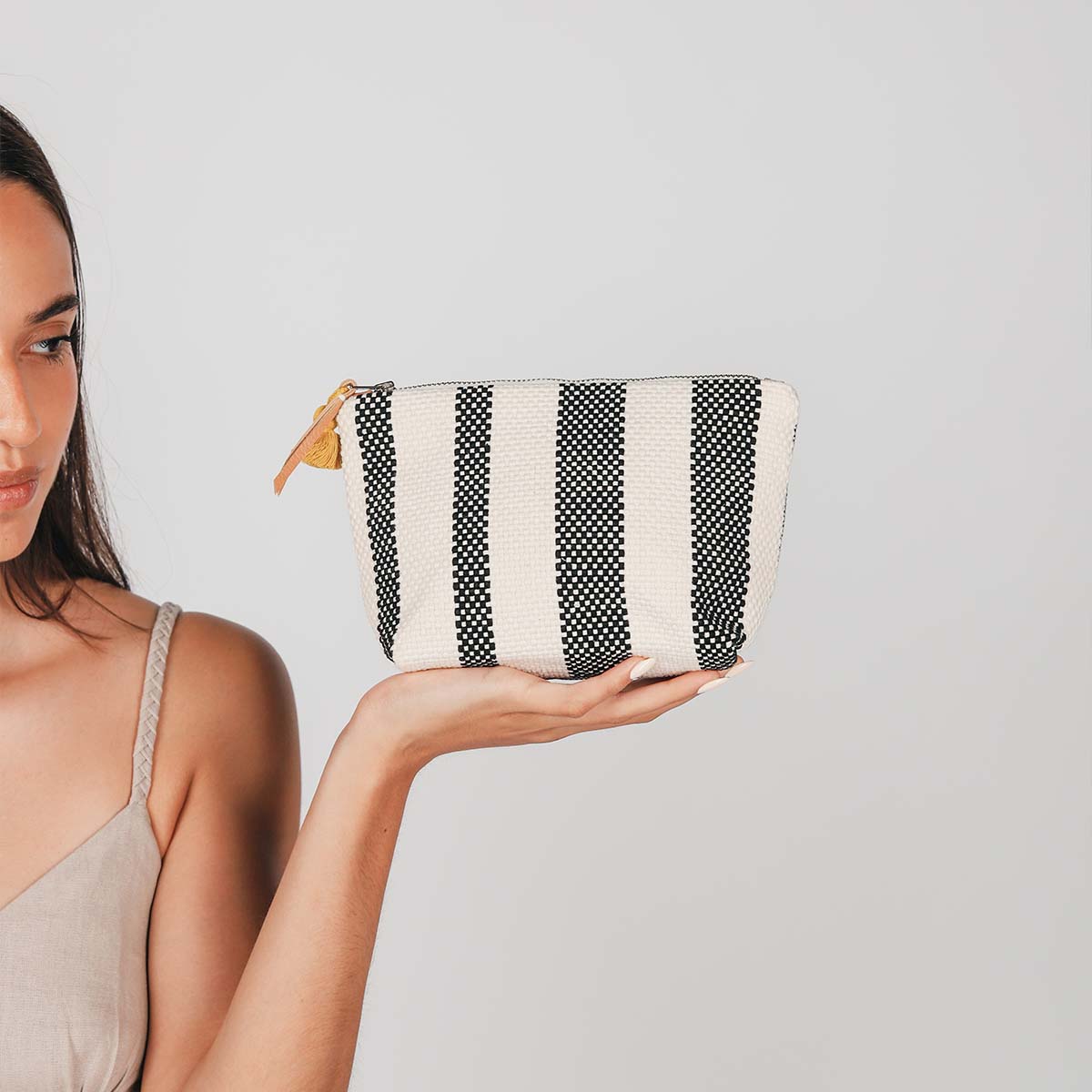 A model holds the hand woven artisan Mini Cristina Cosmetic Pouch in Tourmaline pattern. It shows the front in vertical black and white woven stripes.