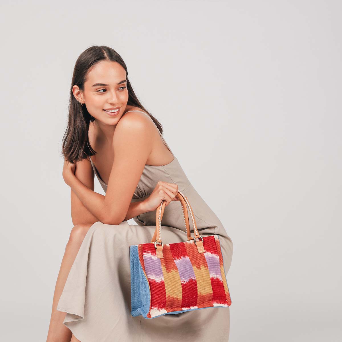 A model sits and holds the hand woven artisan Mini Irma tote in Raspberry Paleta. 