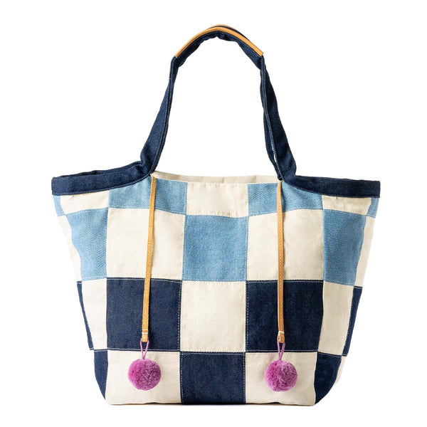 The front of the hand woven artisan Rosa Tote in 90s Denim. The 90's Denim has dark blue and light blue checkerboard squares. It has pink pompoms attached to leather cords.