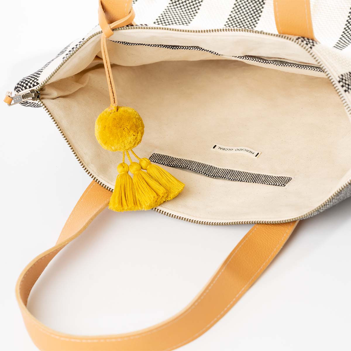 A zoomed-in image of the interior of the Angela Tote in Tourmaline pattern. It has grey and white vertical stripes, leather handles, and a yellow pompom-tassel combination. The interior has a beige fabric color block and a woven black and white hem.