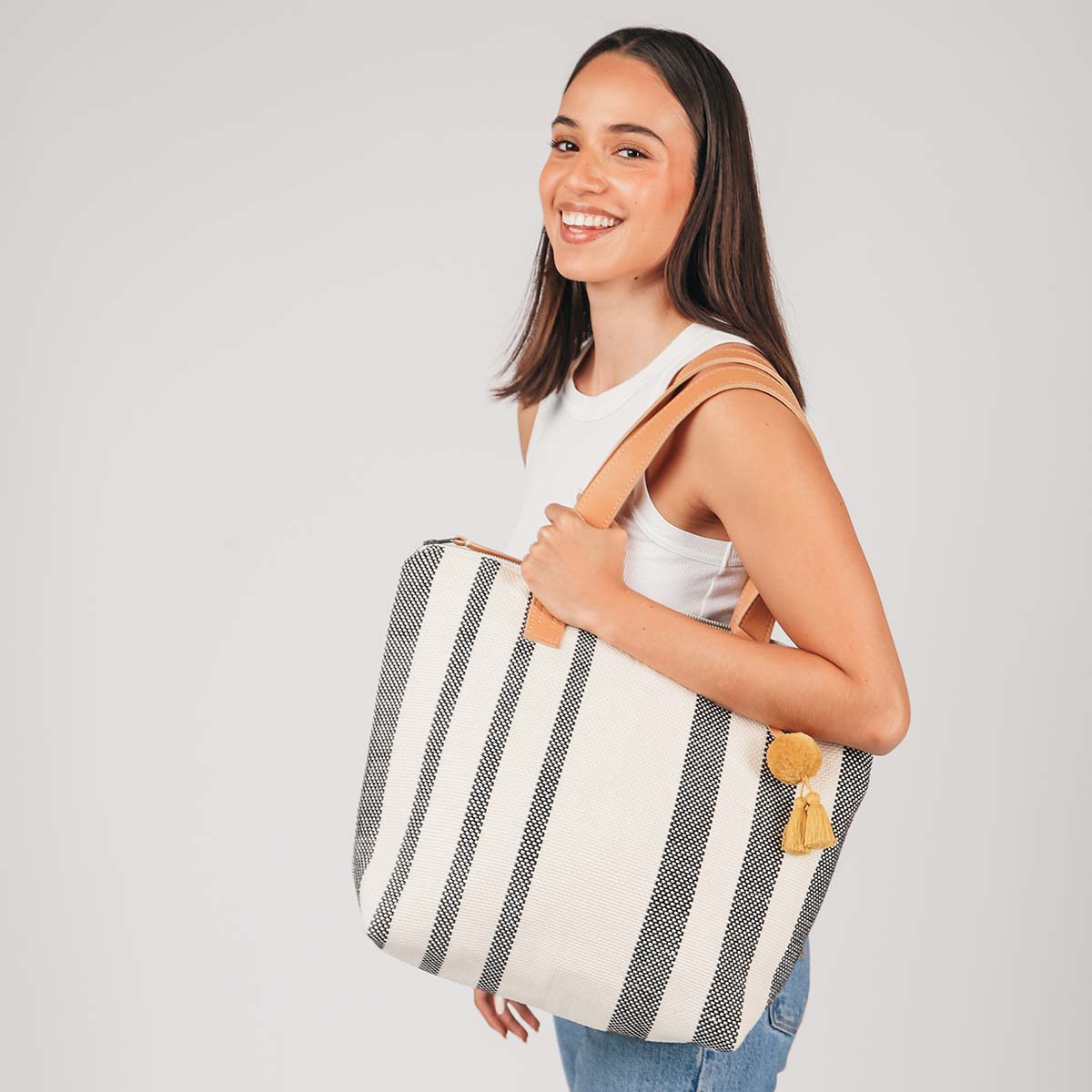 A model holds the Angela Tote in Tourmaline pattern over her shoulder. It has grey and white vertical stripes, leather handles, and a yellow pompom-tassel combination. 