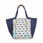 Artisan Rosa Handwoven Tote in Denim Jaspe. The center has an abstract blue square pattern over a white background. The sides have a dark denim wash fabric. The handles have leather detailing. It has two mint green tassels attached to leather cords.